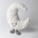 A crescent moon plush toy with a soft white furry texture, embroidered black eyes and a smile, and small gray legs, photographed against a plain light gray background.