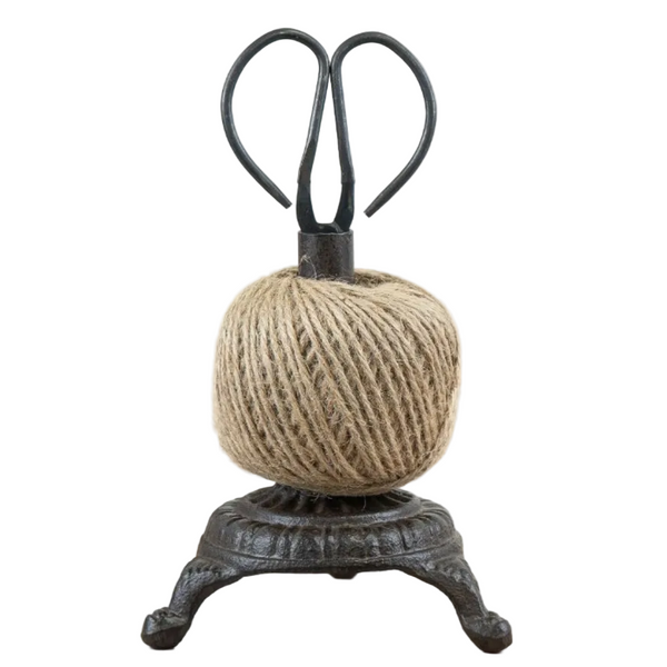 A decorative black metal scissors stand holding a ball of natural jute twine, set on a light wood surface against a plain white background.