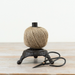 A ball of natural jute twine wound around a small black metal spool, placed on a light wood surface with a pair of black-handled scissors in front, against a plain off-white background.
