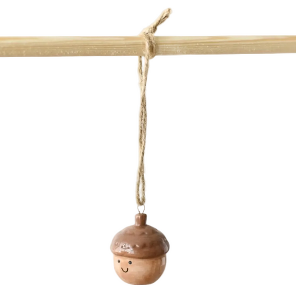 A small wooden acorn ornament with a smiling face hangs from a natural twine string tied to a light wood rod against a plain white background.