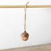 A small wooden acorn ornament with a smiling face hangs from a natural twine string tied to a light wood rod against a plain white background.