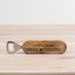 A wooden bottle opener engraved with 'OFFICIAL BEER TESTER' and a star, resting on a light wood surface against a white background.
