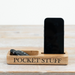 A rectangular wooden desk organizer labeled 'POCKET STUFF' with one open compartment holding car keys and a black smartphone standing in the upright slot, shown on a light wood surface against a white background.