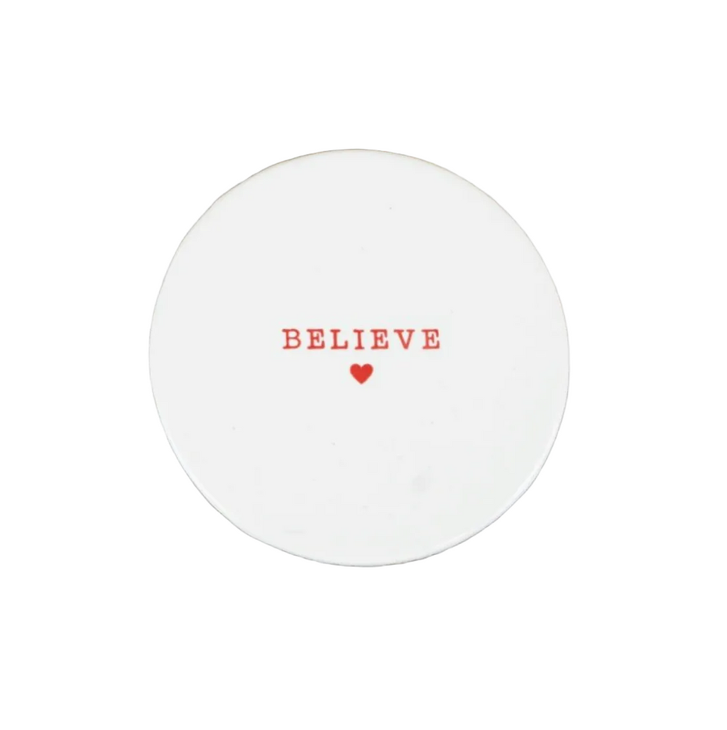 A round white ceramic coaster with the word 'BELIEVE' printed in red serif letters and a small red heart underneath, placed on a rustic white surface.