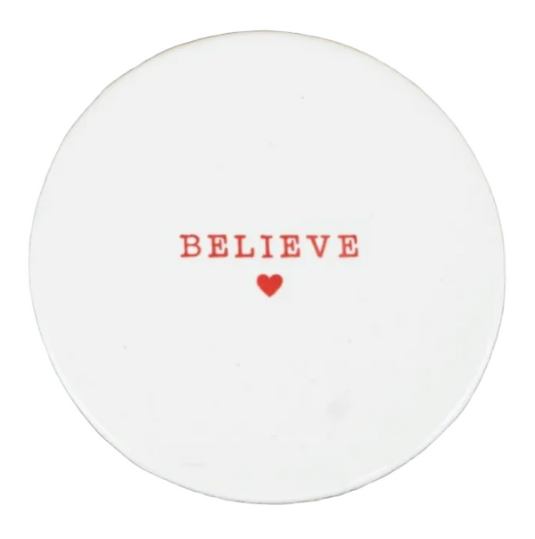 A round white ceramic coaster with the word 'BELIEVE' printed in red serif letters and a small red heart underneath, placed on a rustic white surface.