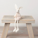 A white felt rabbit doll wearing a cream dress with small pink floral print, sitting on a rustic light wood stool against a soft off-white wall.