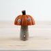 A small decorative mushroom-shaped house figurine with an orange-brown pumpkin cap, a tan wooden-looking stem and body, white window details, and a small star on the door, standing on a light wood surface against a pale background.