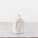 A small white ceramic house-shaped candle holder with gold-toned cutout details, including a crescent moon, stars, and two Christmas trees, displayed on a light wooden surface against a white background.