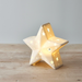 A five-pointed star-shaped ceramic night light with star-shaped cutouts, glowing with a warm yellow light from within, set on a light wooden surface against a neutral gray background.