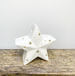 A white ceramic star-shaped candle holder with gold star cutouts, displayed on a rustic wooden surface against a plain light gray wall.