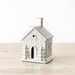 A small ceramic house-shaped candle holder or ornament with a textured off-white and beige brick-like pattern, standing on a light wooden surface against a plain white background.