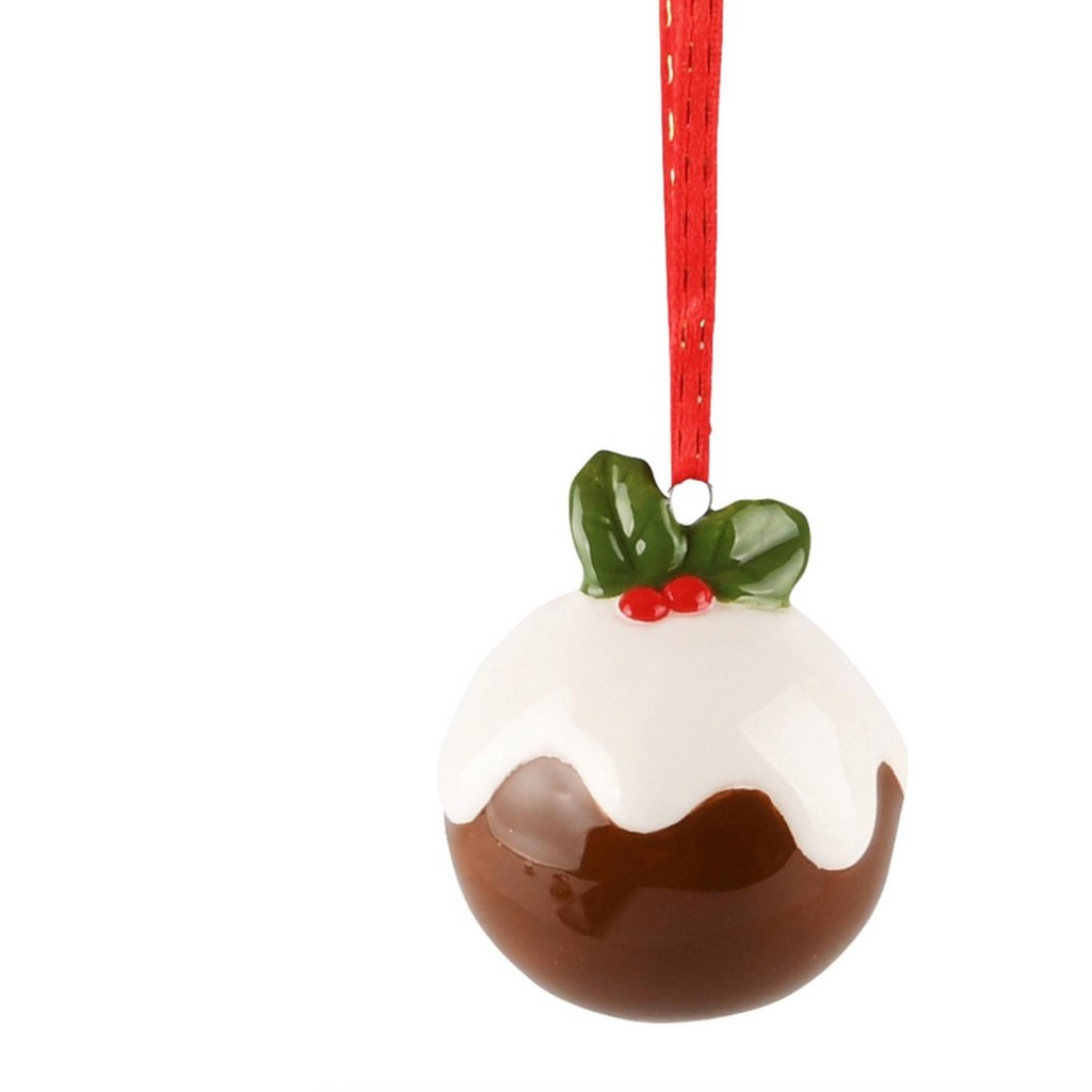 A Christmas pudding ornament shaped like a glossy brown pudding with white icing, a green holly leaf, and red berries, hanging from a red ribbon on a white background.