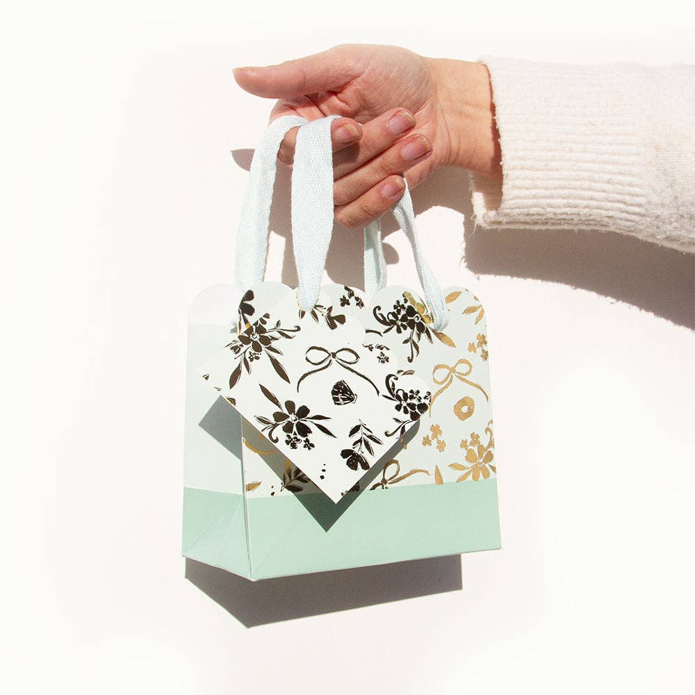 A hand holding a small gift bag with white handles, featuring a cream body with black floral patterns and a mint green base, set against a plain white background.