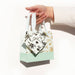 A hand holding a small gift bag with white handles, featuring a cream body with black floral patterns and a mint green base, set against a plain white background.