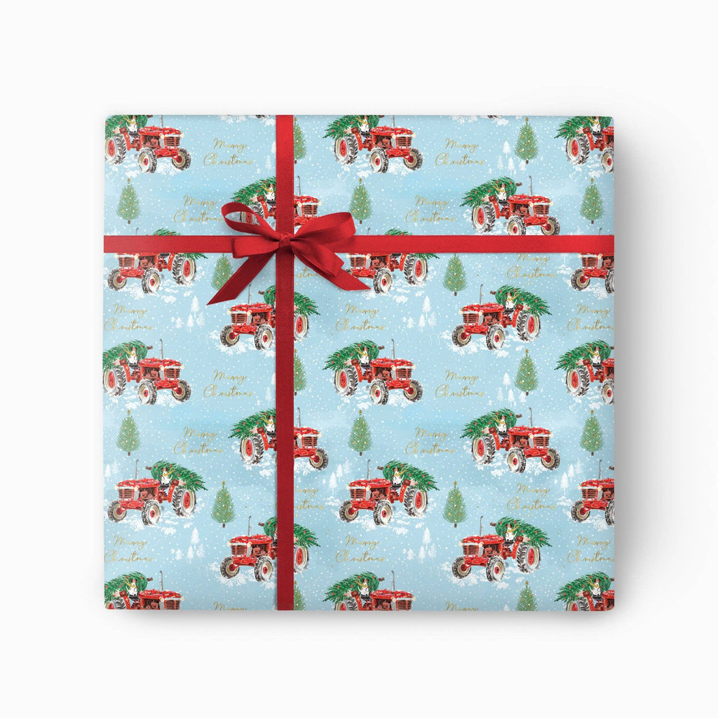 A square gift box wrapped in light blue Christmas-themed wrapping paper with a red tractor and tree pattern, tied with a red ribbon and bow on a white background.