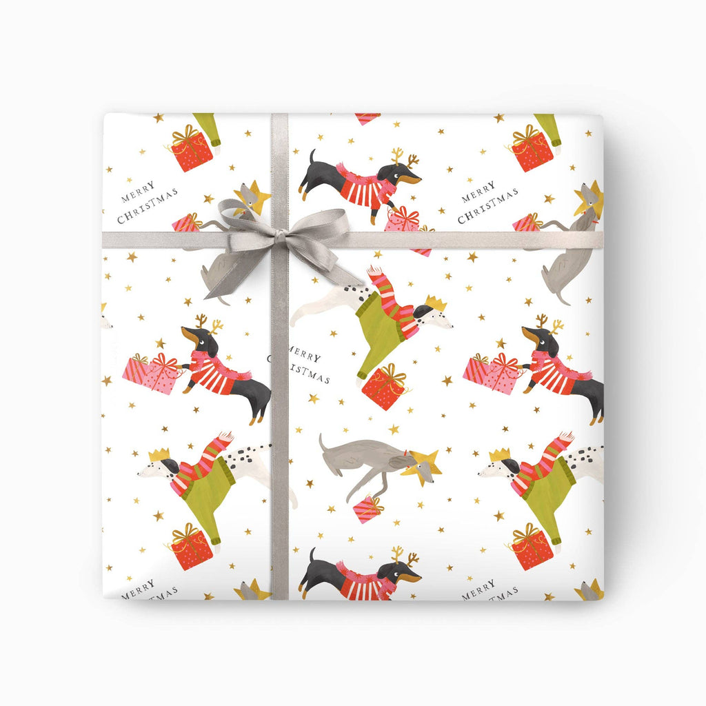 A square gift box wrapped in white Christmas-themed paper with a pattern of dachshunds in red sweaters, green boots, red and pink presents, gold stars, and 'MERRY CHRISTMAS' text, tied with a gray ribbon and bow.