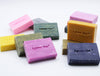 A group of handmade rectangular bars of soap in various colors including pink, green, yellow, purple, and blue, arranged on a plain white background. Each bar is embossed with the brand name 'Agnes + Cat'.