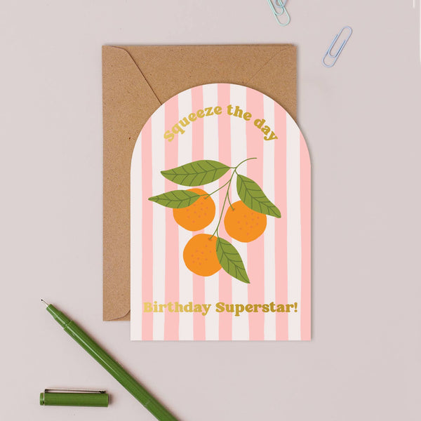 Greeting card with oranges and text 'Squeeze the day, Birthday Superstar!' on a striped background, with an envelope and pen.